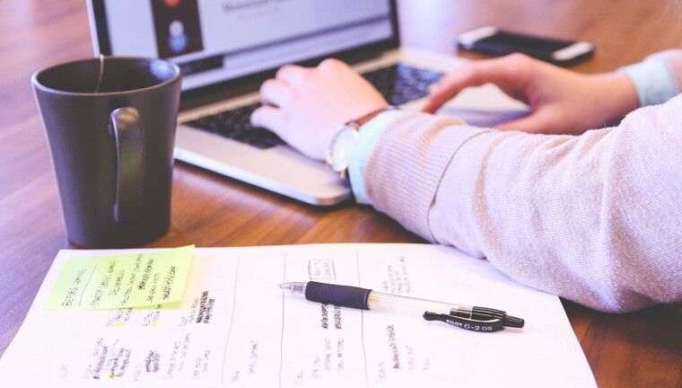 Alt-Text: Das Foto zeigt die Hände einer Frau, die auf ihrem Laptop tippt, eine Tasse und ein Blatt Papier.